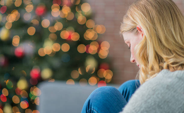 A Caucasian woman is indoors in her living room. There is a Christmas tree in the background. The woman is wearing warm clothing. She is sitting on the couch and looking sad because she is alone on Christmas day.