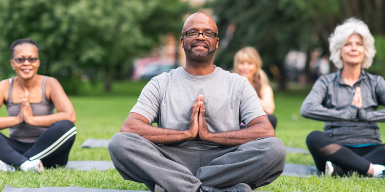 A multi-ethnic group of seniors is attending a yoga class outdoors. The group is sitting on yoga mats. They are meditating. The individual in focus is a black man. He is sitting at the front of the group. He is smiling directly at the camera.
