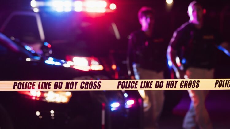 Two multi-ethnic police officers standing in front of patrol cars, behind police tape, wearing bulletproof vests and duty belts. The policewoman is a mature African-American woman in her 40s. Her partner is a mid adult man in his 30s. It is nighttime and the emergency lights on top of the vehicles are flashing.  The focus is on the cordon tape in the foreground.