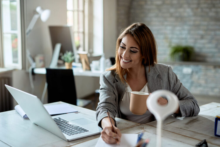 Young happy female entrepreneur using computer and taking notes while drinking coffee in the office.